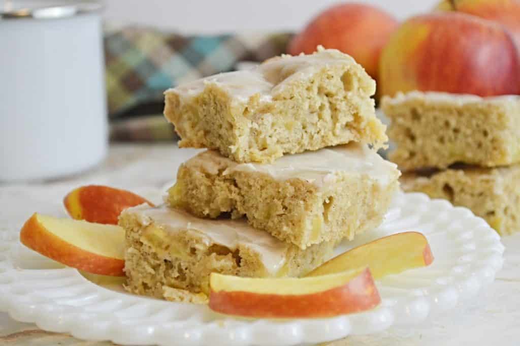 Three glazed apple bars stacked on a white plate, surrounded by sliced apples.
