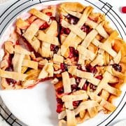 A lattice-topped pie with visible red and pink filling, possibly cherry or berry, is placed on a cooling rack. Sliced apples and whole cherries are nearby on a white surface.
