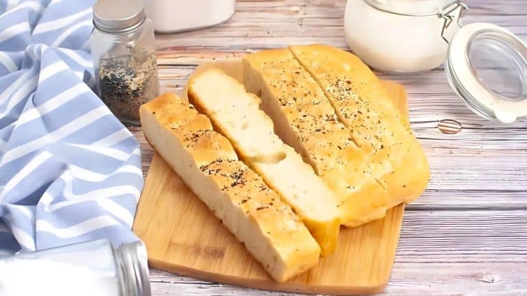 Sliced focaccia bread with herbs on a wooden board, next to a blue-and-white striped cloth and glass jars on a wooden surface.