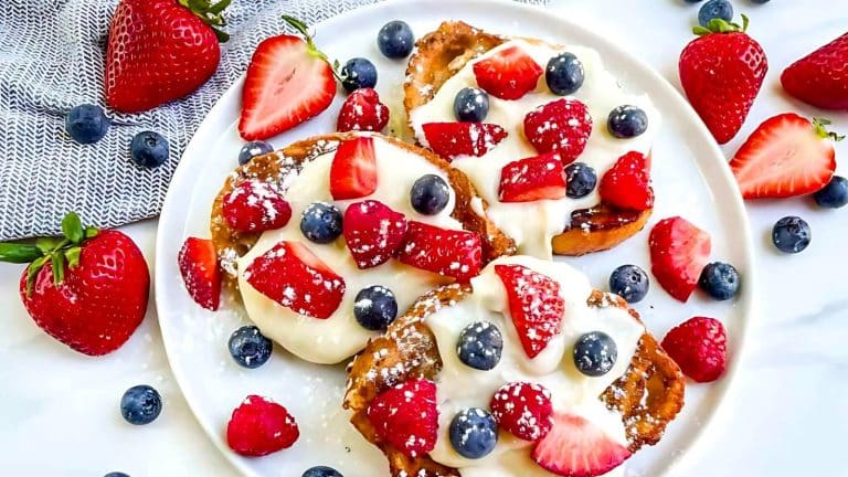 A plate of French toast topped with whipped cream, sliced strawberries, blueberries, and a dusting of powdered sugar, surrounded by additional strawberries and blueberries.