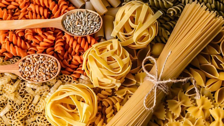 An assortment of uncooked pasta varieties and seeds arranged with wooden spoons, including fusilli, tagliatelle, penne, and bow-tie pasta tied with string.