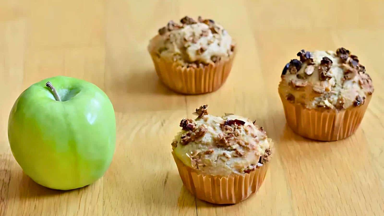 A green apple is placed on a wooden surface next to three muffins topped with granola.