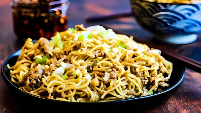 A plate of noodles mixed with chopped vegetables and meat, served with chopsticks. A bowl and jar are in the background on a wooden table.