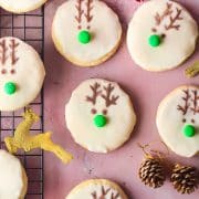 Cookies decorated with icing and reindeer designs are placed on a cooling rack with festive decorations, including a small wreath, pinecones, red berries, and gold reindeer figures.