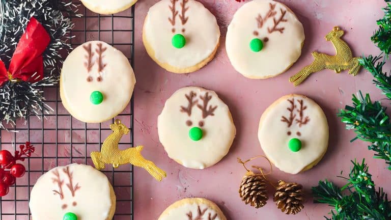 Cookies decorated with icing and reindeer designs are placed on a cooling rack with festive decorations, including a small wreath, pinecones, red berries, and gold reindeer figures.