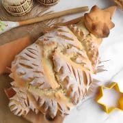 A decorative loaf of bread shaped like a tree on a wooden board, surrounded by a spoon, a star-shaped cookie cutter, and stalks of wheat on a marble surface.