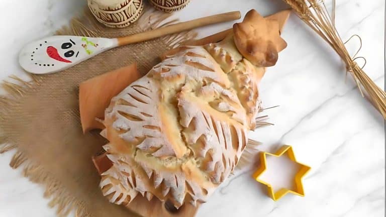 A decorative loaf of bread shaped like a tree on a wooden board, surrounded by a spoon, a star-shaped cookie cutter, and stalks of wheat on a marble surface.