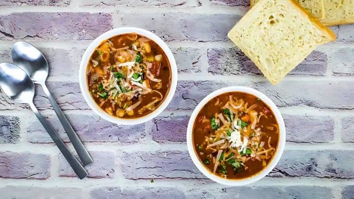 Two bowls of vegetable soup with grated cheese on top, placed on a textured surface. Two slices of bread and metal spoons are on the side.