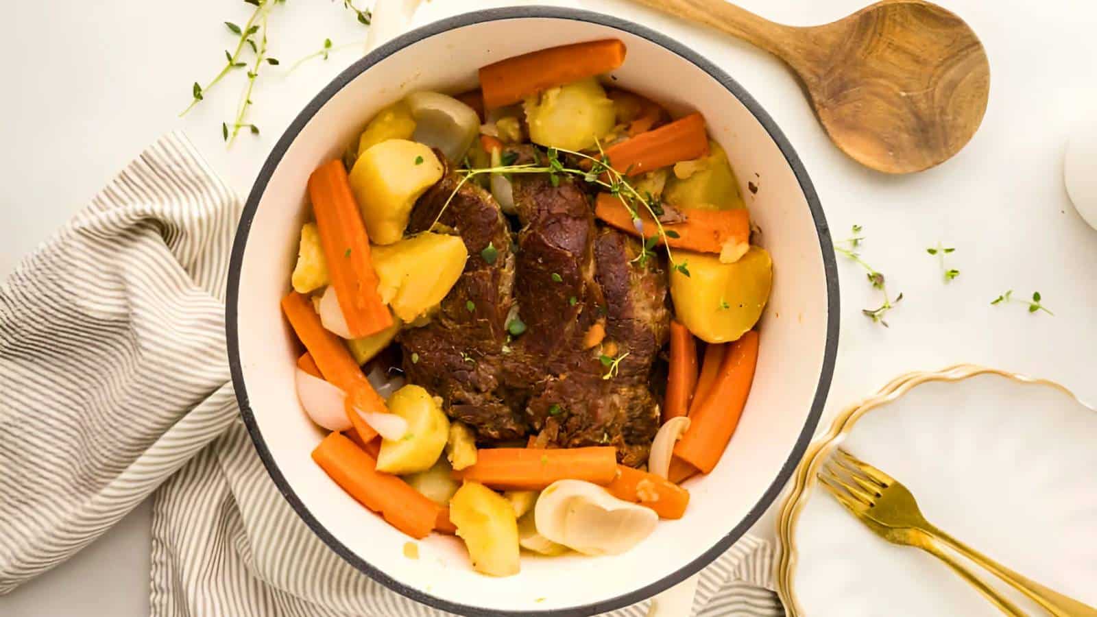 A pot roast with beef, carrots, potatoes, and herbs in a white pot. A wooden spoon and a striped cloth are nearby, along with a plate and fork.