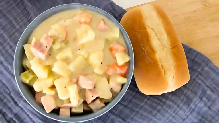 A bowl of creamy potato and vegetable stew with diced ham, accompanied by a plain bread roll on a textured cloth.