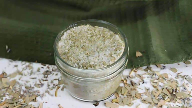 A small glass jar filled with a mixture of coarse salt and herbs sits on a white surface, surrounded by scattered herb flakes. A green cloth is partially visible in the background.
