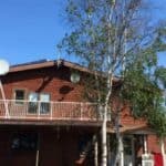 A two-story brown off grid home with a white balcony, satellite dishes, solar panels on the roof, and trees in the front yard under a clear blue sky.
