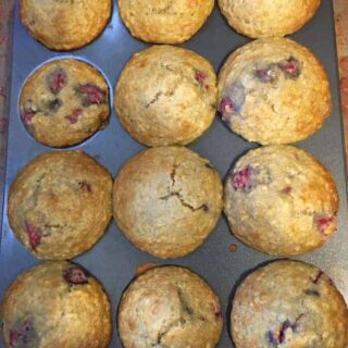 A muffin tin holds twelve golden-brown muffins, some with visible berry pieces—one compartment is empty, hinting at a popular recipe for raspberry lemon almond muffins.