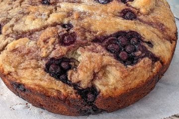 A close-up of a round baked cake with a golden crust and visible clusters of blueberries throughout.