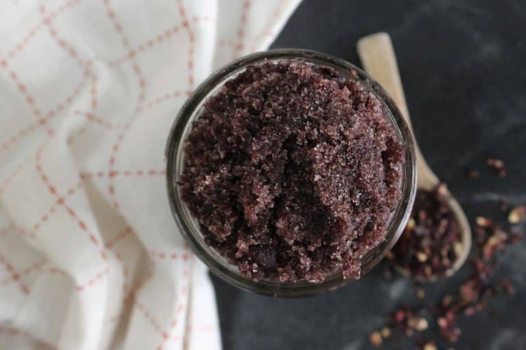 A glass jar filled with dark purple hibiscus DIY sugar scrub sits on a black surface next to a wooden spoon and a white cloth with red stitching.