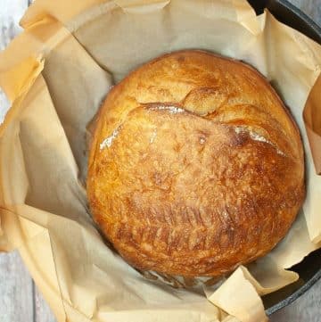 A round loaf of golden brown bread sits on parchment paper inside a black cast iron pot.