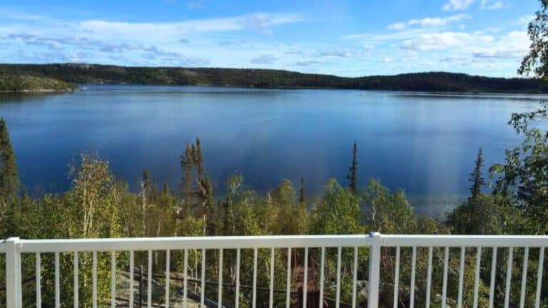A clear lake surrounded by trees and hills is viewed from behind a white metal railing on a sunny day with a blue sky and scattered clouds.