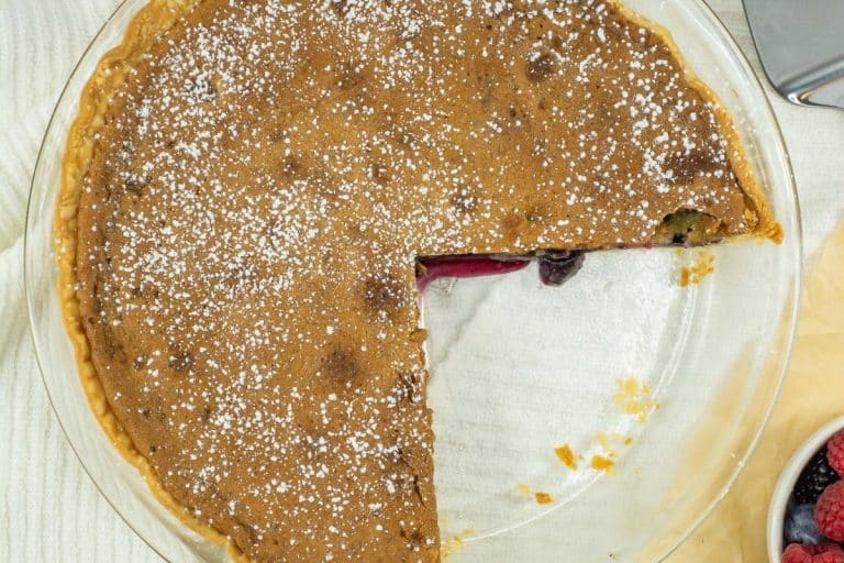 A partially eaten pie with a golden-brown top, dusted with powdered sugar, sits in a clear glass dish—a beautiful recipe idea for Thanksgiving recipes—its missing slice revealing the vibrant red fruit filling inside.