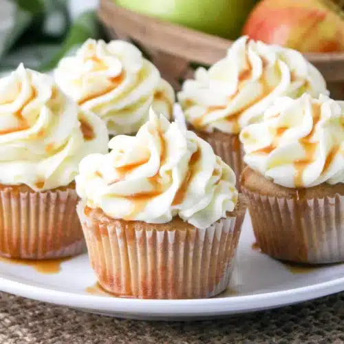 Four cupcakes with white frosting and caramel drizzle are arranged on a white plate, with apples in a basket behind them—perfect inspiration for autumn apple desserts.