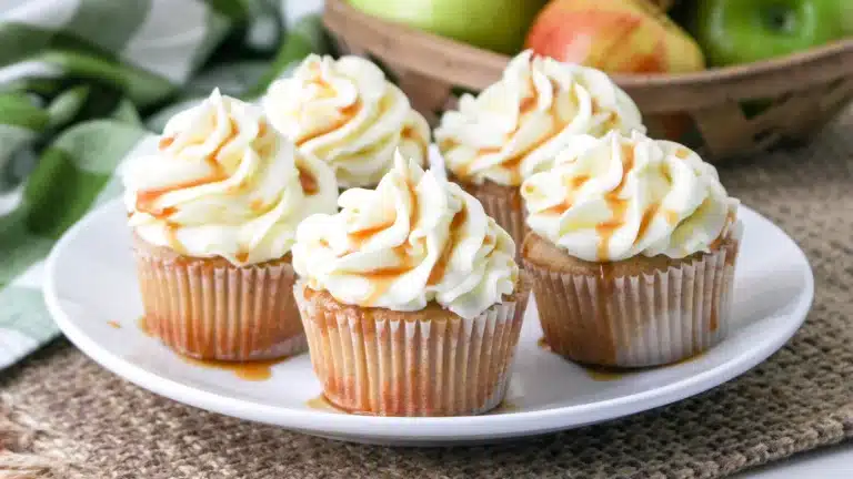Four cupcakes with white frosting and caramel drizzle are arranged on a white plate, with apples in a basket behind them—perfect inspiration for autumn apple desserts.