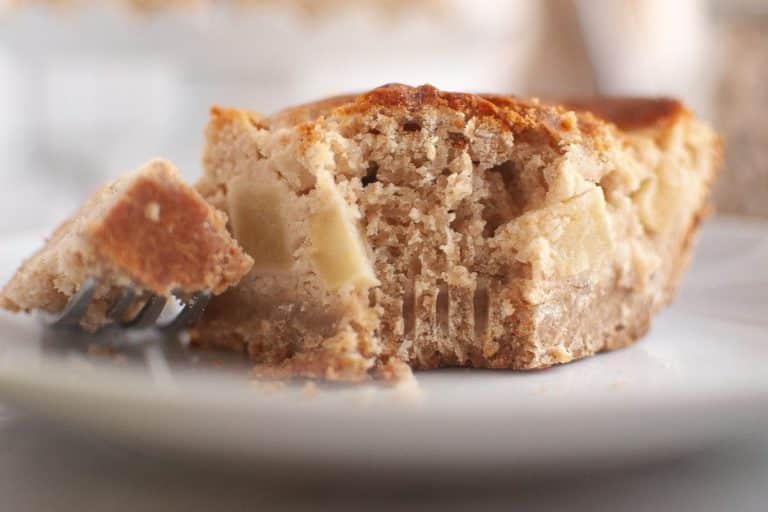 Close-up of a partially eaten slice of apple sour cream coffee cake with visible apple pieces, resting on a white plate with a fork holding a bite-sized piece.