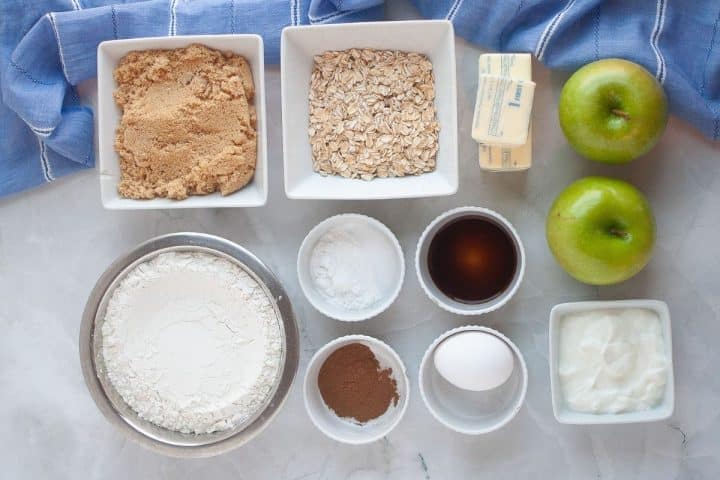 Top-down view of baking ingredients for apple sour cream coffee cake including brown sugar, oats, butter, green apples, flour, cinnamon, baking powder, vanilla extract, an egg, and yogurt on a marble surface.