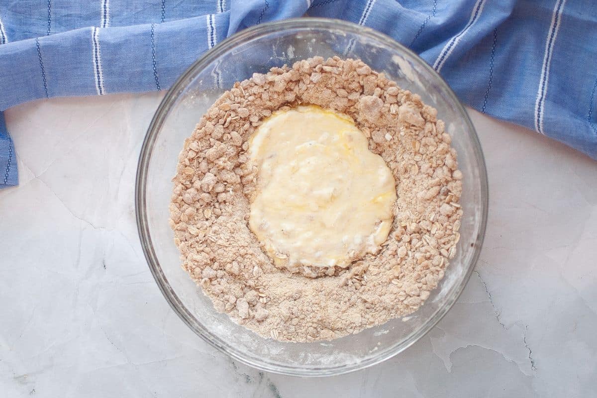 A glass bowl contains a mixture of dry oats and flour with a well in the center filled with wet ingredients, placed on a light countertop beside a blue striped cloth.