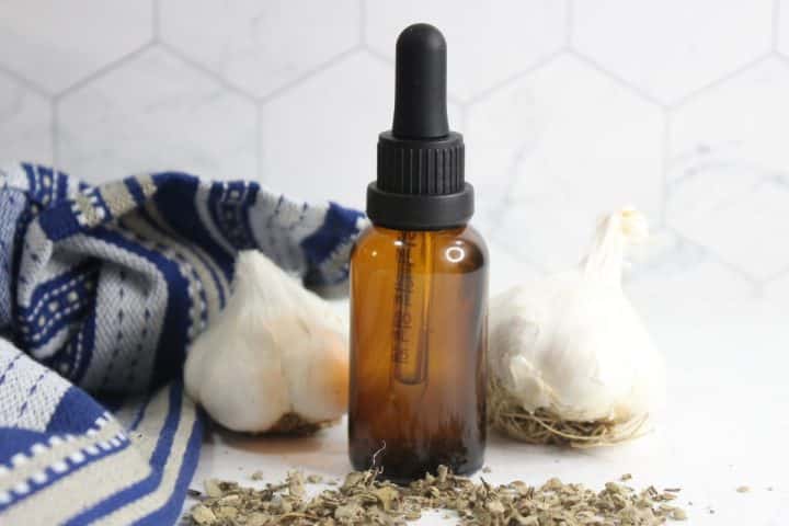 Amber dropper bottle placed between two garlic bulbs, with dried herbs and a blue patterned cloth nearby on a light surface.
