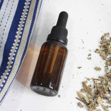 A brown glass dropper bottle of mullein garlic ear oil sits on a white surface, accompanied by a blue and white striped fabric and a small pile of dried herbs.