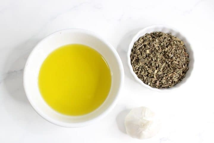 A bowl of olive oil, a bowl of dried herbs, and a whole garlic bulb on a white surface.