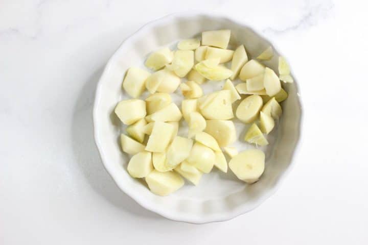 A round white dish containing several peeled and chopped garlic cloves on a white surface.
