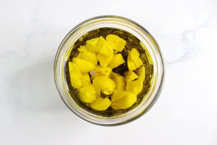 A glass jar filled with olive oil and yellow garlic cloves, photographed from above on a white surface.