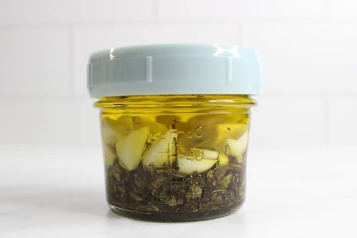 A small glass jar with a light blue lid contains garlic cloves and herbs submerged in oil, set against a white tiled background.