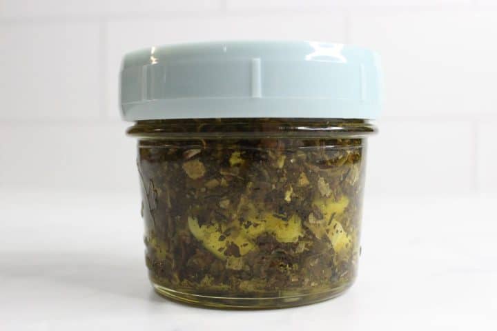 A small glass jar with a light blue lid, filled with a mixture of chopped herbs and oil, sits on a white surface against a white tiled background.
