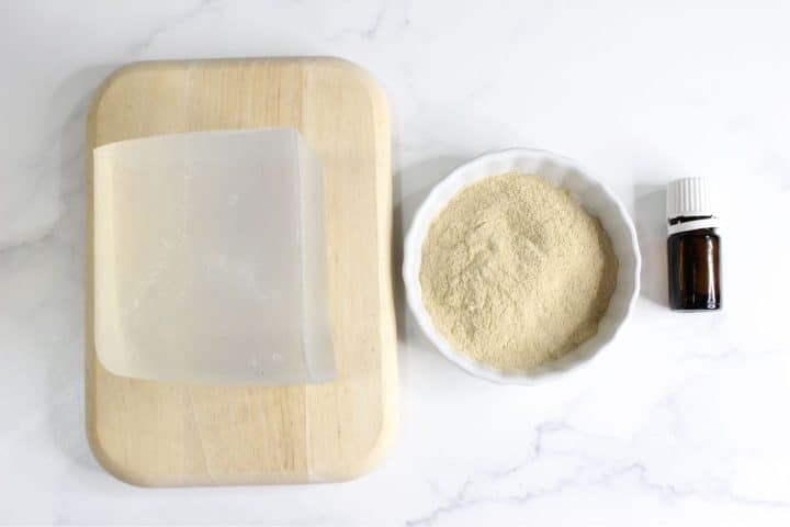 A rectangular block of clear soap base on a wooden board, a white bowl of beige powder, and a small amber glass bottle on a white surface.
