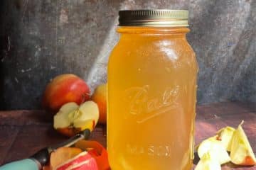 A mason jar filled with golden homemade apple cider vinegar sits on a wooden surface, surrounded by whole and sliced apples and a corer.