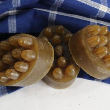Three round brown massage soaps with raised nubs rest on a white surface beside a blue and white checkered cloth, perfect for trying a homemade lemon soap recipe.