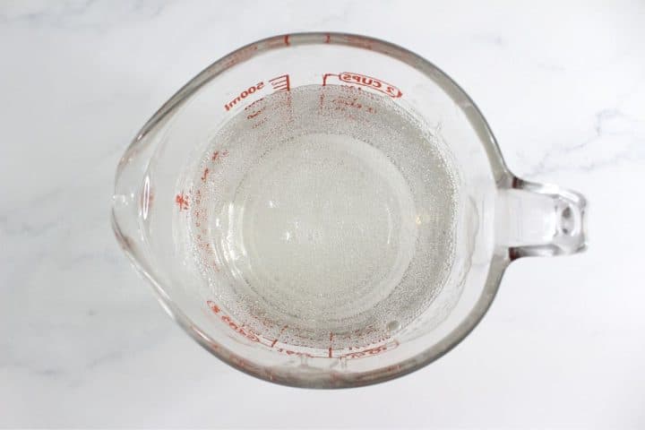 A glass measuring cup filled with a clear liquid, marked with red measurement lines, placed on a white marble surface.