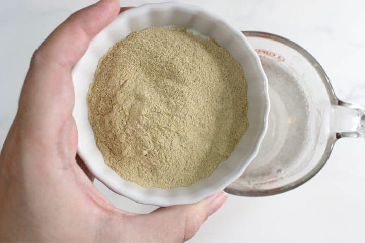 A hand holding a white bowl filled with dry yeast above a glass measuring cup containing water.