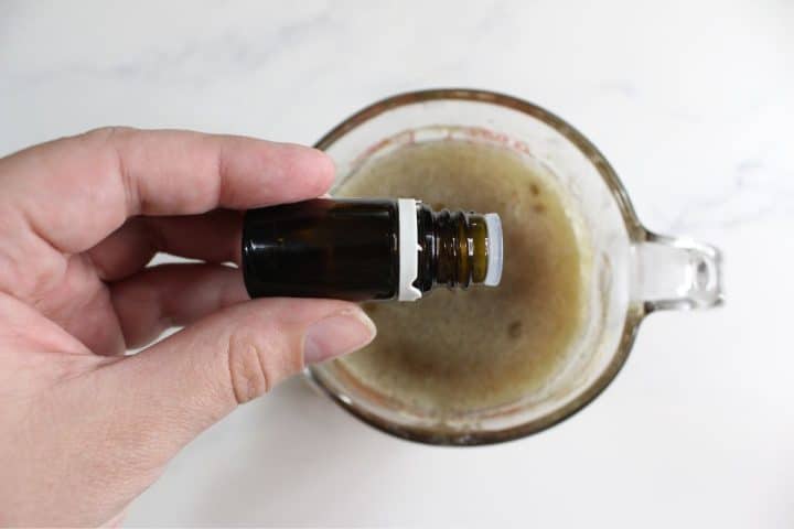 A hand holds a small brown glass bottle over a measuring cup containing a frothy brown liquid on a white surface.