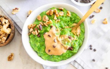 A bowl of bright green oatmeal makes for a filling, cold-weather breakfast, topped with peanut butter, chopped walnuts, and chocolate chips, with a gold spoon beside it.