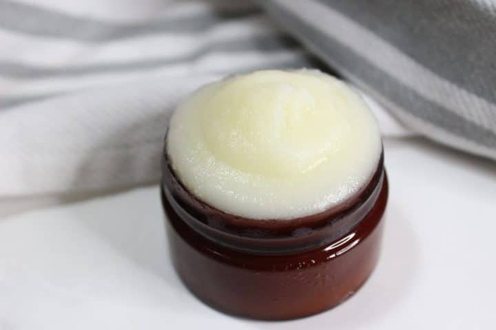 A small brown jar filled with rose beef tallow face cream, placed on a white surface with a folded striped cloth in the background.