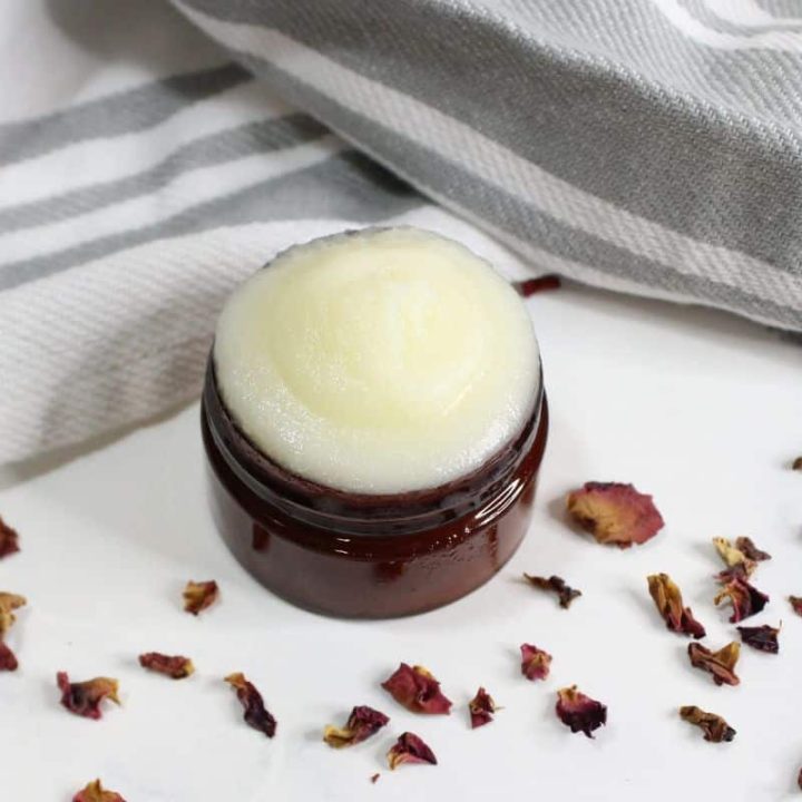 A small brown jar filled with rose petal beef tallow face cream sits on a white surface, surrounded by dried flower petals and a striped cloth.
