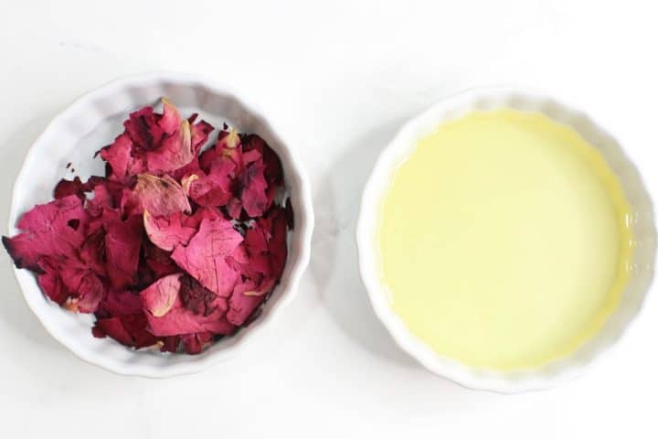 A white dish filled with dried rose petals is next to another white dish containing a pale yellow liquid, both placed on a white surface.