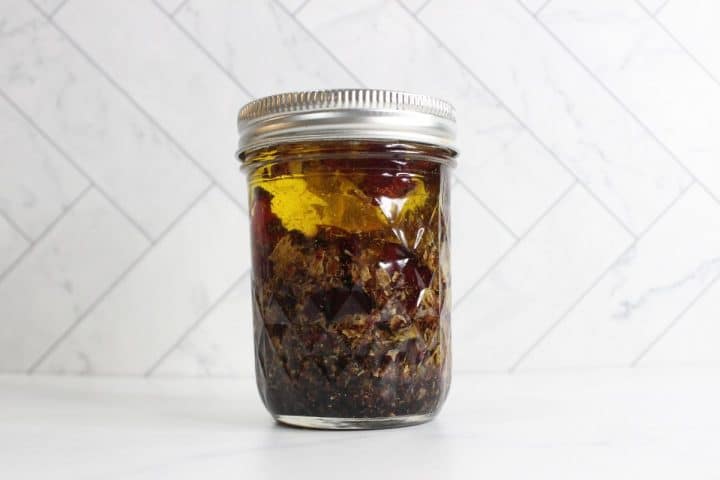 A glass jar with a metal lid containing a mixture of chopped ingredients and oil, placed on a white surface with a tiled background.