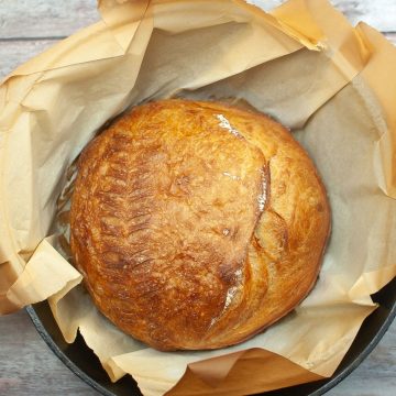 A round loaf of golden dutch oven sourdough bread sits on parchment paper in a baking dish, one of many recipes for food off the grid.