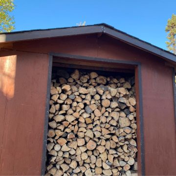A neatly stacked pile of firewood fills the open entrance of a brown wooden shed under a clear blue sky.
