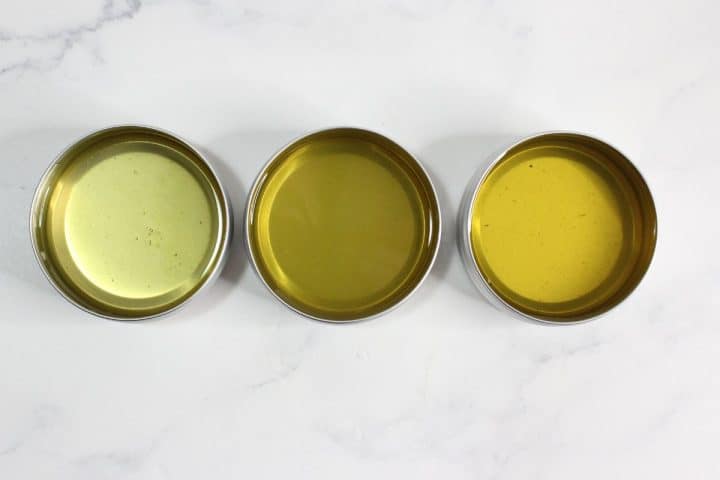 Three small metal tins filled with different shades of yellow-green liquid, arranged in a row on a white marble surface.