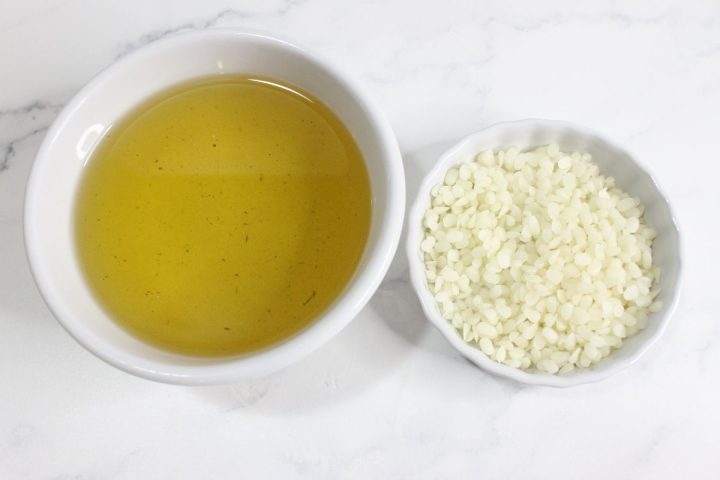 A bowl of yellow liquid next to a small bowl filled with white wax pellets on a white marble surface.