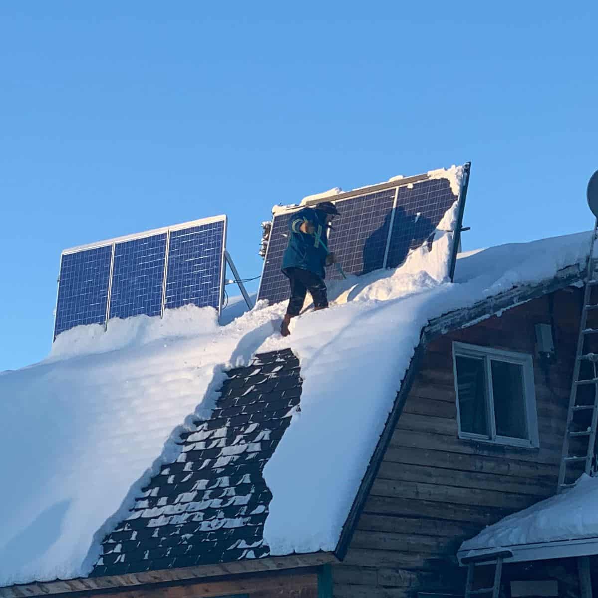 A person stands on a snow-covered roof, clearing snow off solar panels mounted above a wooden house on a clear day.